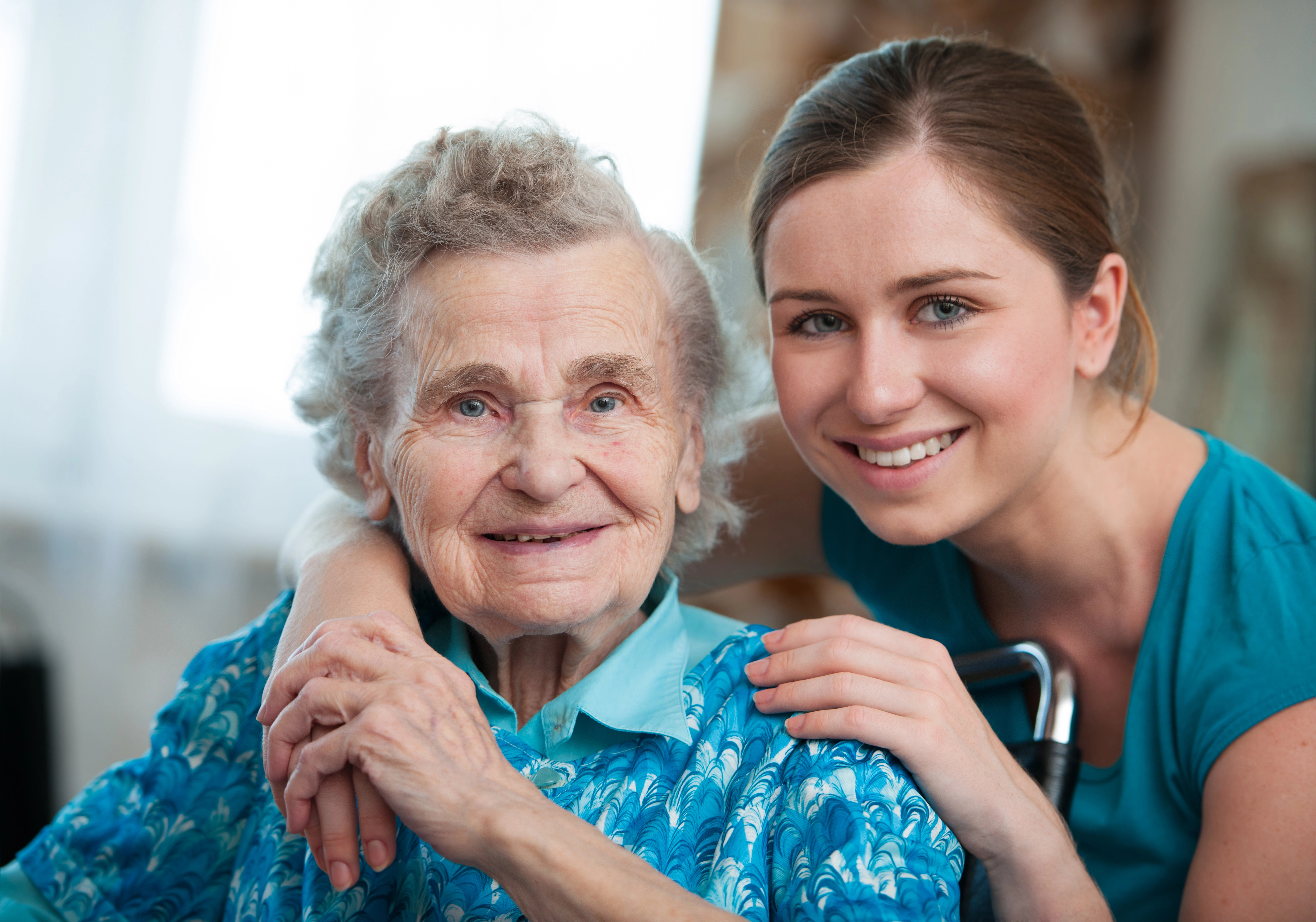 A young woman smiles warmly as she puts her arm around an older woman, both looking directly at the camera. They are both wearing blue shirts and appear to be close.